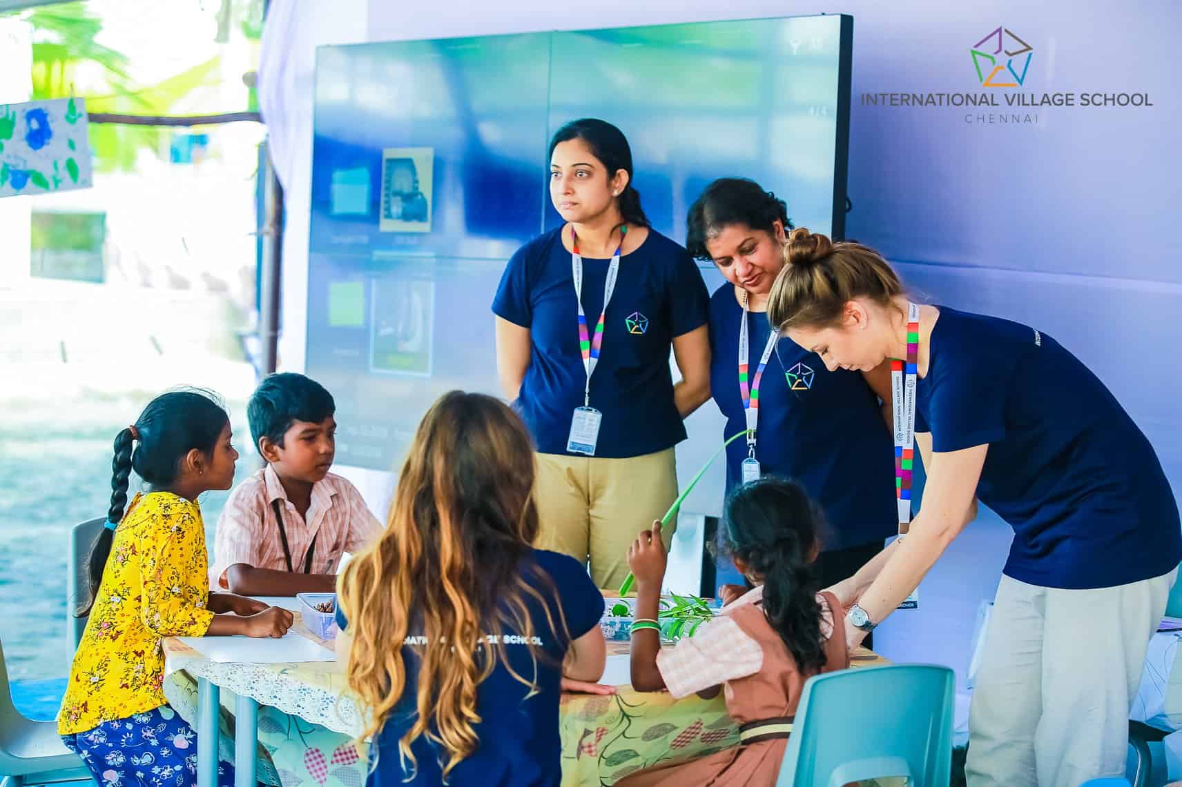 Three students sitting around a table with one girl holding a plant and three IVS teachers standing and guiding them. One IVS teacher sitting with the students.