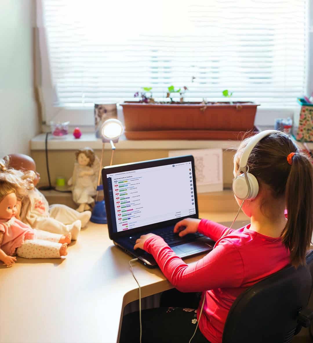 A girl in pink dress wearing headset typing in laptop with few toys beside the laptop  in the table
