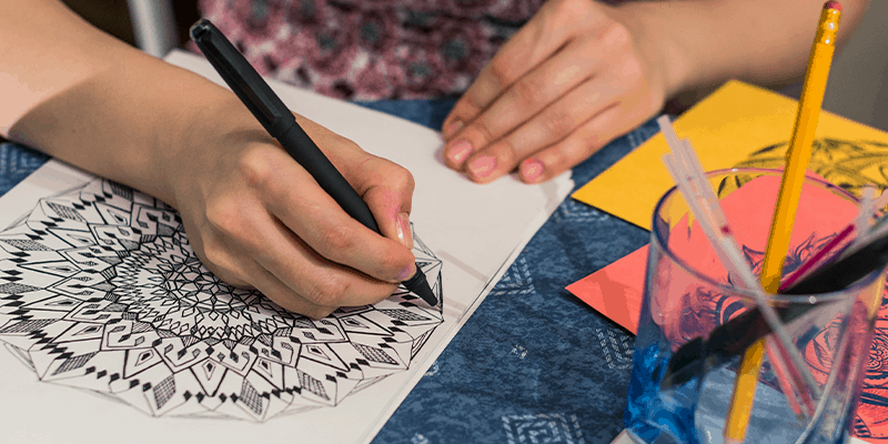 A hand holding black pen and drawing mandala art on a sheet of paper.